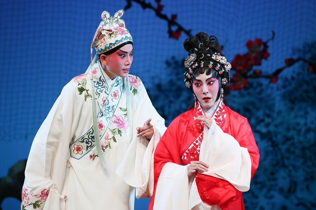 A man and woman in stage makeup and costume, singing. Both wear long robes with floral embroidery and dramatic brows and eyeshadow.