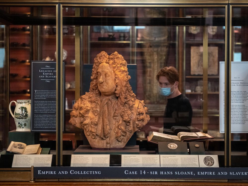 A glass case with the newly positioned bust of Sloane, which is a terracotta color, next to a small sign that has paragraphs of text explaining how he profited from enslavement. Behind, a visitor walks by wearing a mask.
