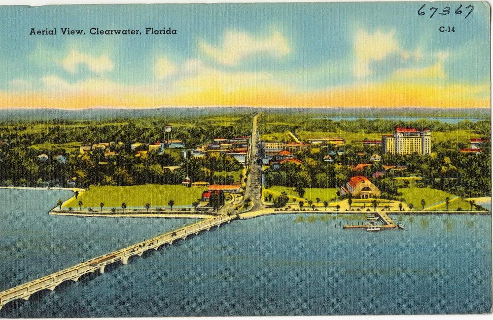 An aerial view of the city on the bay, with blue water in foreground and a bridge extending to the left