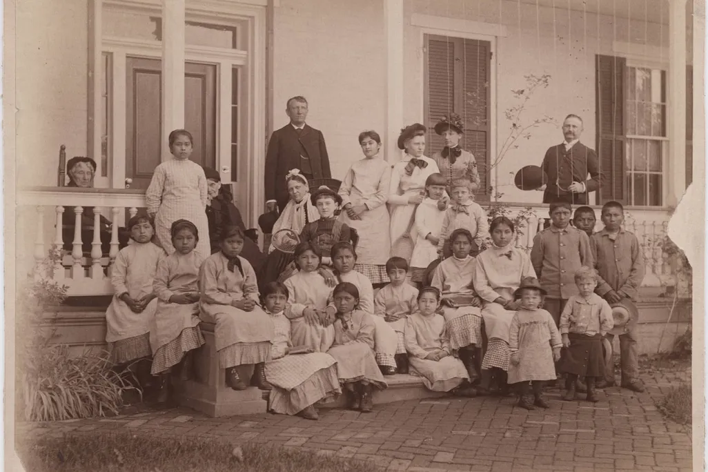 A sepia toned image of a group of young women, with a few young boys and girls, sitting in front of a white house on a porch. Pratt stands in dark garb behind the group