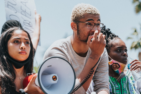 Three young activists are outside at a protest – looking off into the distance. They all appear to be African American. One is holding a bullhorn and has a determined look on her face. Another is holding the bullhorn’s microphone and speaking into it.