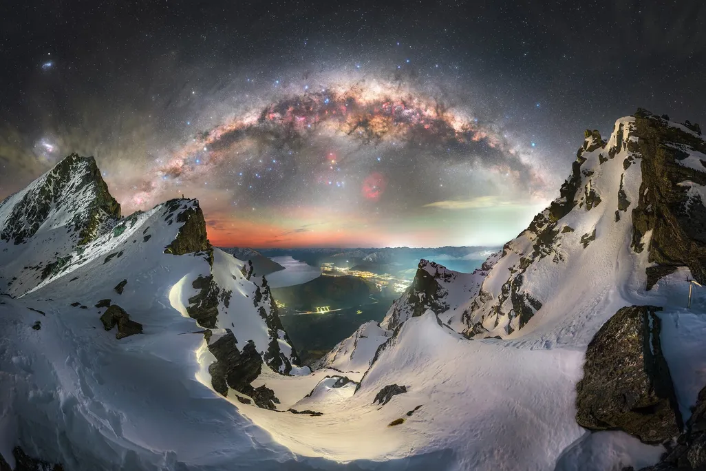 the milky way arched over mountain peaks with an illuminated town in the background