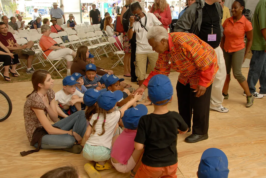 A woman with dark skin and bright orange shirt with African print shakes the hands of young children, all in blue ballcaps, sitting on a wooden dancefloor outside.