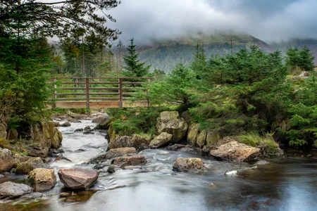 In England&rsquo;s Lake District, a wooden footbridge connects two rocky sides of a riverbank as mountains peek out from the fog in the distance.