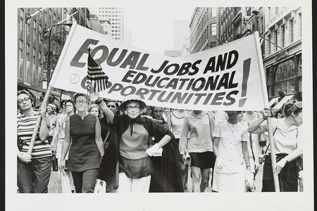 Women marching down the street holding a banner with the message “Equal jobs and educational opportunities!”