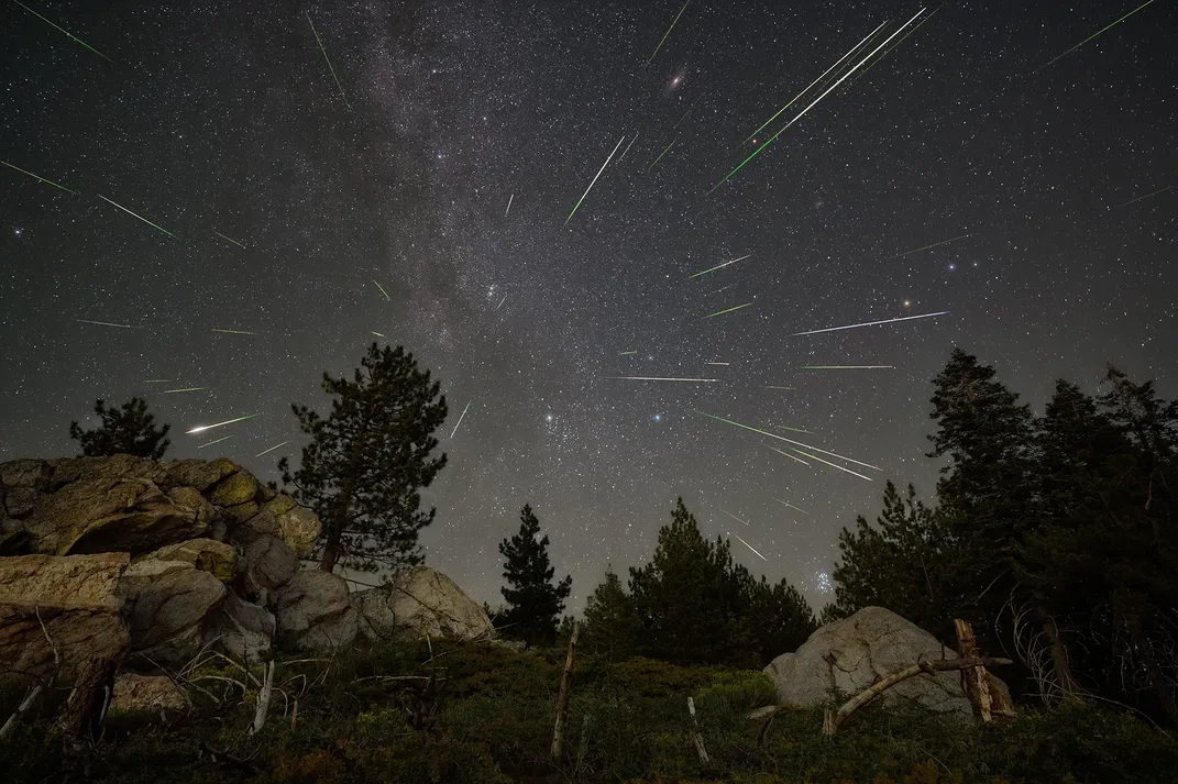 several meteors streak across the sky, seeming to emanate from the center of the frame, with trees and rocks in the foreground