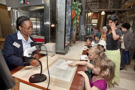 A group of students wait in line at a Post Office. Each student has a stamped envelope they are handing to the postal worker behind the counter.