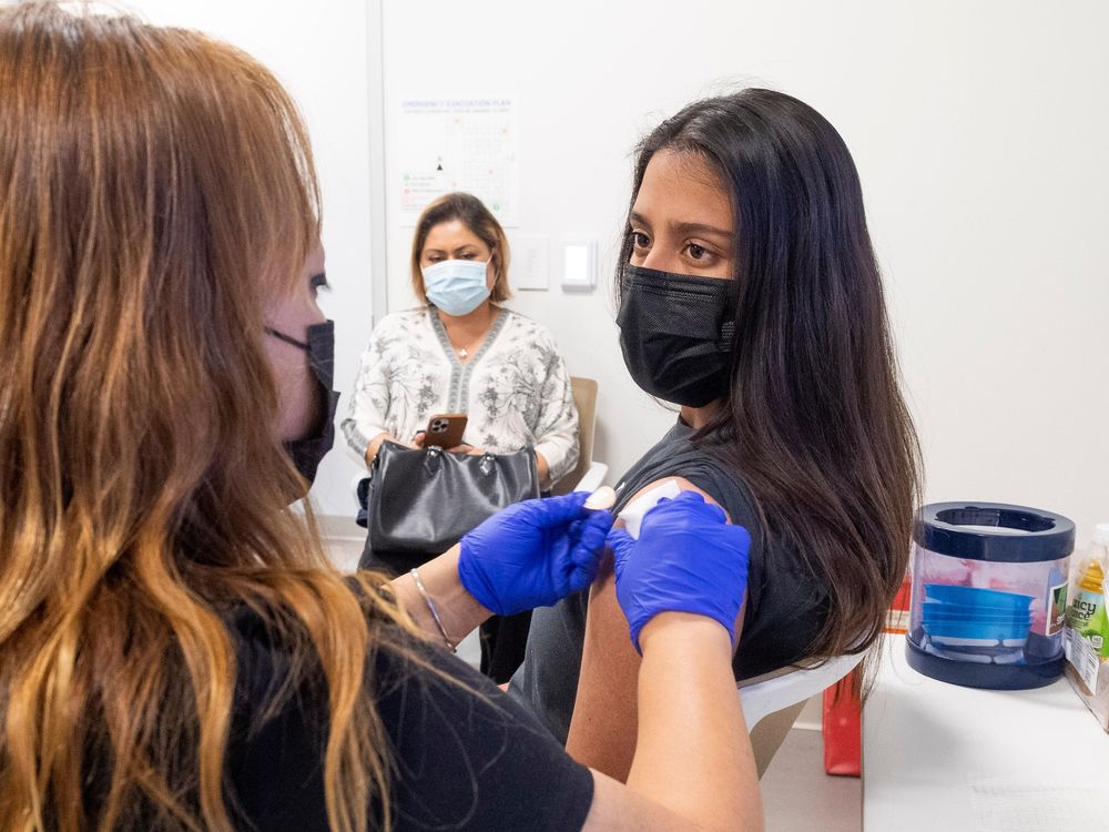 The back of a nurse's head is out of focus on the left side of the image. To the right, a teen girl sits with her sleeve rolled up facing the nurse as she places a bandage on the teen's shoulder.