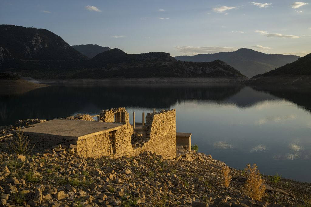 ruins of a building stand out from the side of a hill against a low lake with mountains in the background
