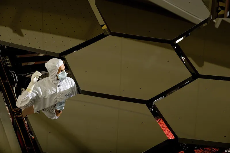 a man in white gloves, clothes, hood and mask stands amid six large hexagonal mirrors