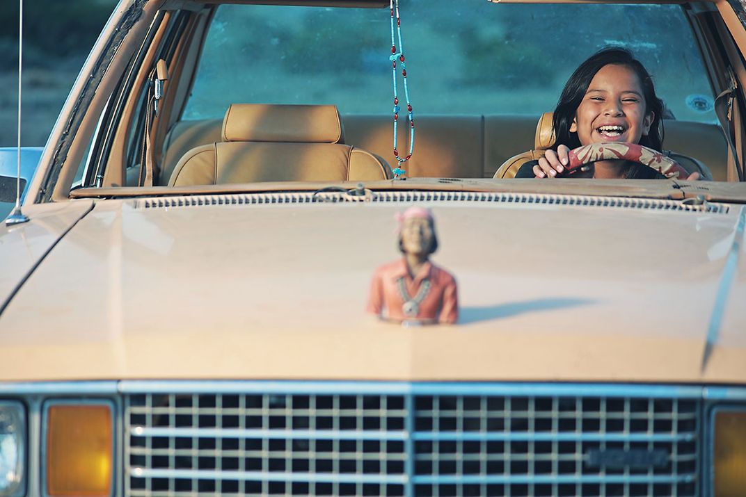 A kid sits behind the wheel of a tan car, laughing, with a Native American figure as a hood ornament.