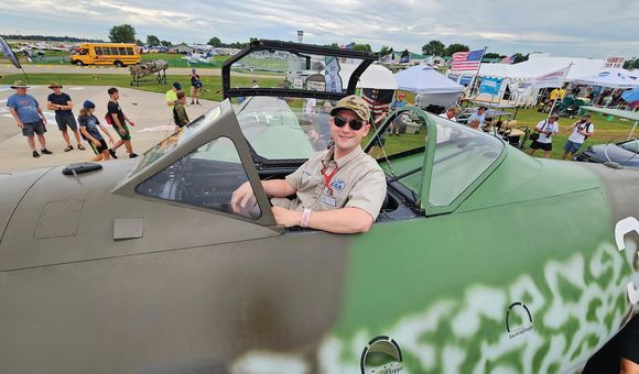 A white man in his 30s sits in the cockpit of a World War II-era military airplane. He is smiling and wearing sunglasses and a ballcap.