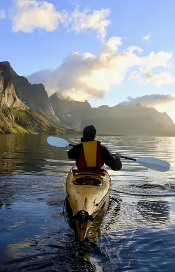 Active Journeys - a view from behind of a person kayaking towards a mountain in the distance with the sun peeking through clouds just over the horizon.