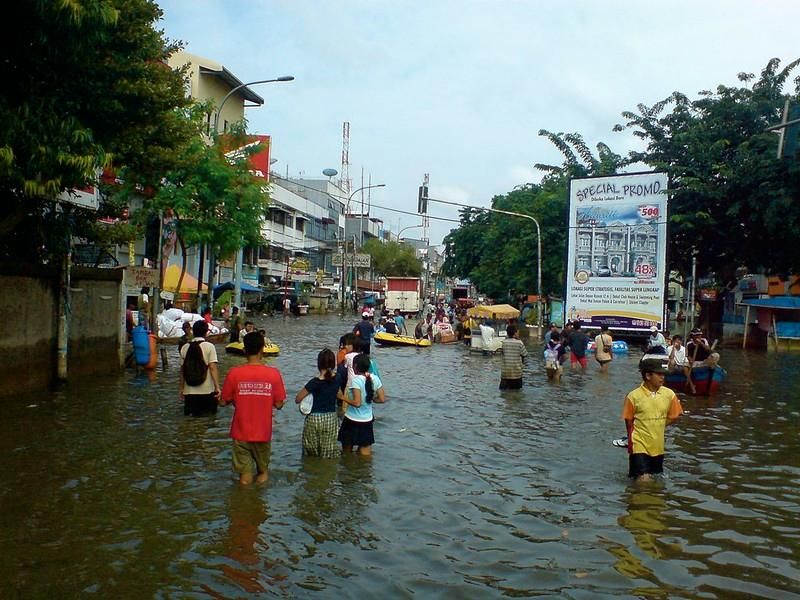 A view of a flooded marketplace in Jakarta. People are wading through knee-high water in the middle of a street. It looks like a typical, bustling street except that the street is entirely flooded.