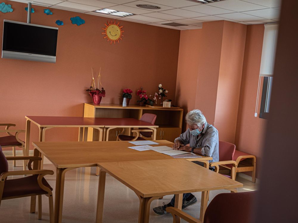 A person sits at a table wearing a mask and writing. The room in the nursing home is otherwise empty and has coral-colored walls