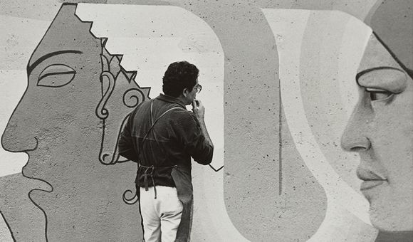 Grayscale image of Emanuel Martínez in a dark colored shirt and apron and light coloroed pants pondering a wall mural with two large faces.