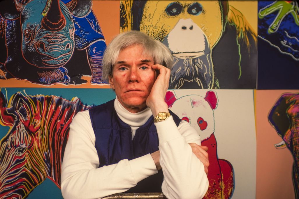 A color image of Warhol, wearing a floppy white wig, a white turtleneck and blue vest, poses with his head on his hand in front of screen printed works featuring rhinos, zebras, and pandas