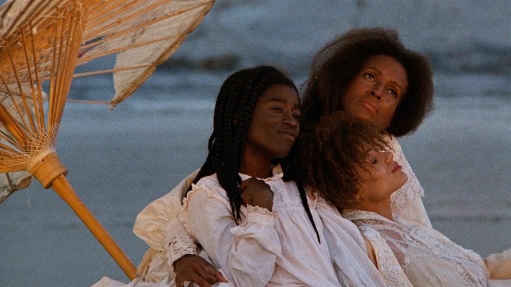 A still photograph from a movie with three females sitting next to each other under an umbrella.