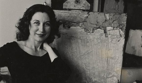 Grayscale photograph of smiling woman dressed in a black top and pants with her right hand resting on her hip and her right hand next to her neck standing in front of an abstract painting on an easel.