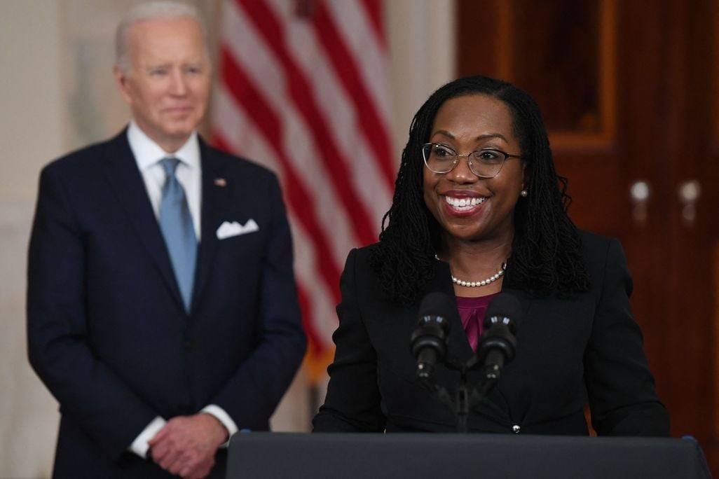 Judge Jackson stands at a podium and smiles, wearing glasses, a string of pearls, a purple top and a black blazer. In the background, President Biden smiles and stands in front of an American flag