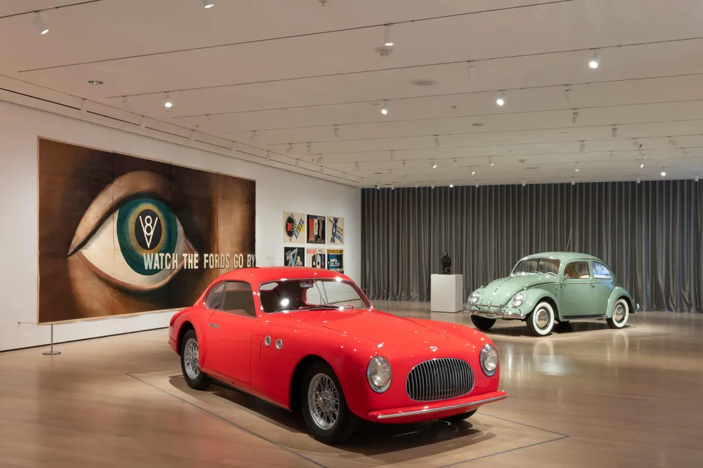 A view of a museum gallery with a bright red car on display in front of a light green Beetle; on the wall, an enormous lithograph of a human eye with the words Watch the Fords Go By