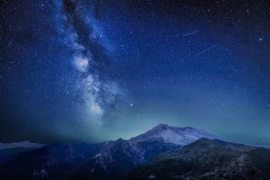 The Milky Way glitters over a blue-green sky full of shooting stars, all overlooking a white-capped mountain