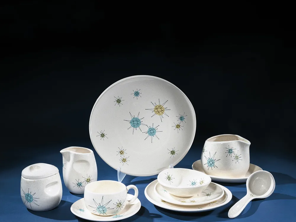 Photographed against a dark blue background is a studio image of a set of cream-colored ceramic dishware bearing a pattern of star-shapes in blue, yellow, and green. There are several pieces, including a platter, a gravy boat with a ladle, a creamer and s