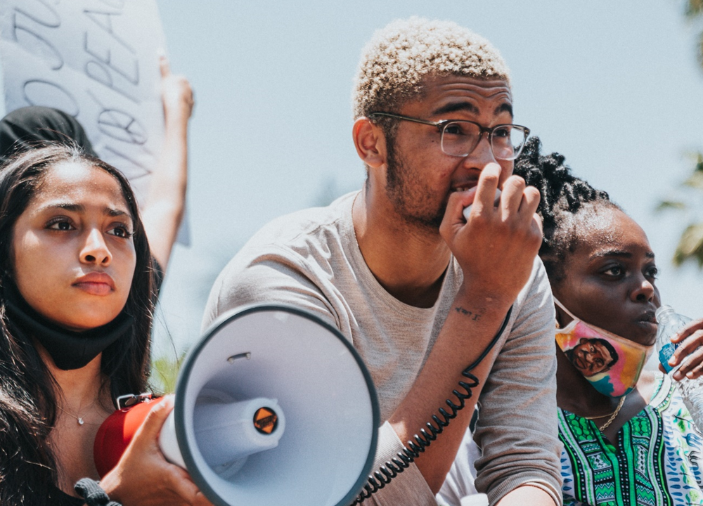 Three young activists are outside at a protest – looking off into the distance. They all appear to be African American. One is holding a bullhorn and has a determined look on her face. Another is holding the bullhorn’s microphone and speaking into it.