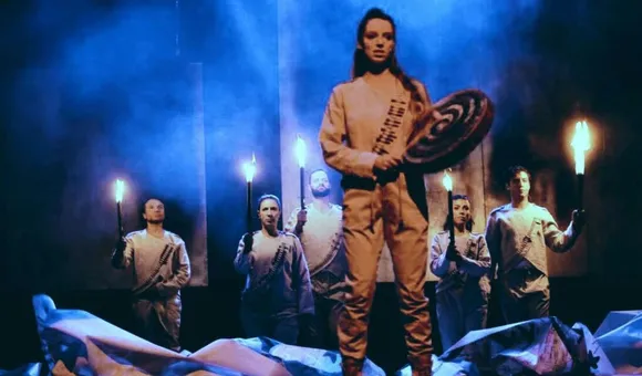 A woman stands on stage, holding a hand drum and a beater stick. Five actors behind her hold up torches. They all wearing matching white outfits with mock bandoliers. The stage is foggy and lit blue.