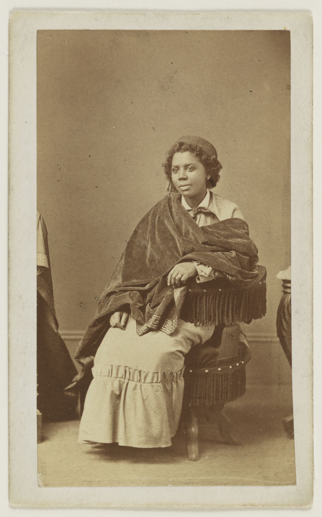 Edmonia Lewis, a Black woman with curled hair wearing a shawl and a dress, sits and looks into the distance in a sepia-toned portrait image