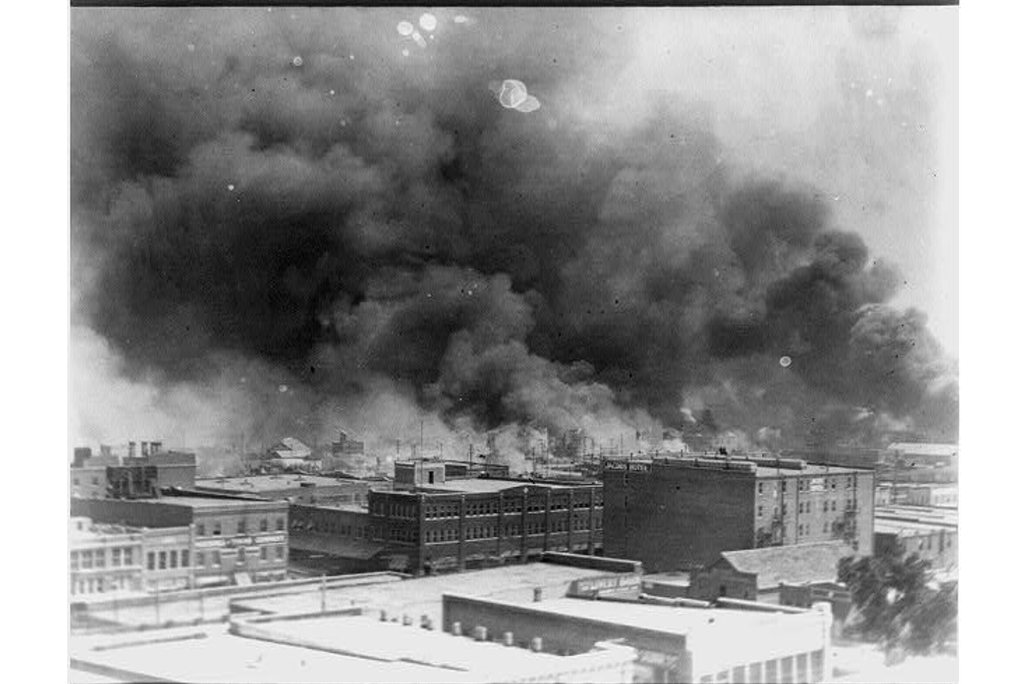 A black and white photo of buildings, from an aerial perspective, with thick plumes of black smoke rising above and obscuring the sky