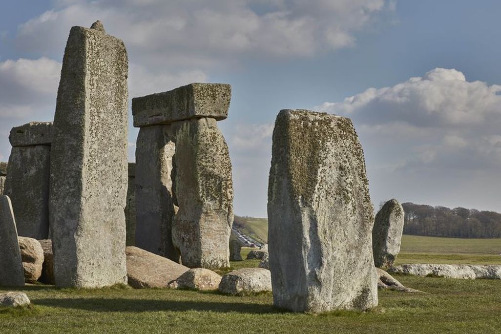 A view of the iconic rocks of Stonehenge on green grass, with a sloping hill visible between two stones in the distance and a line of traffic
