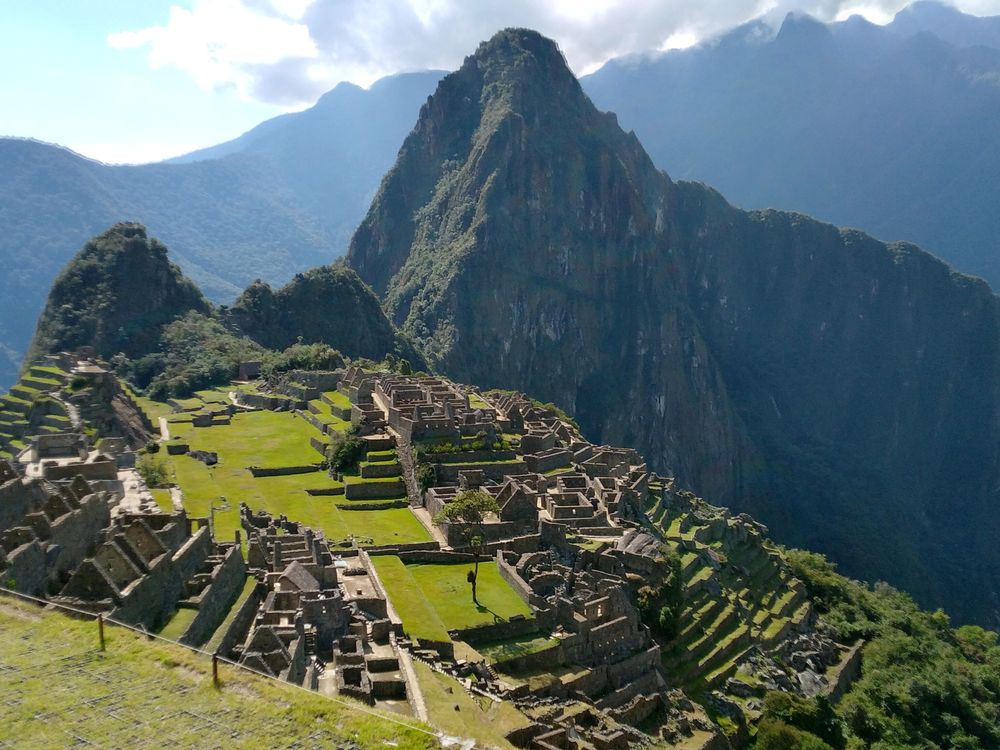 A panoramic view of Machu Picchu from above, with green slopes and angular mountain ridge in distance and the ruins of settlements along a sloping green mountainside; devoid of tourists