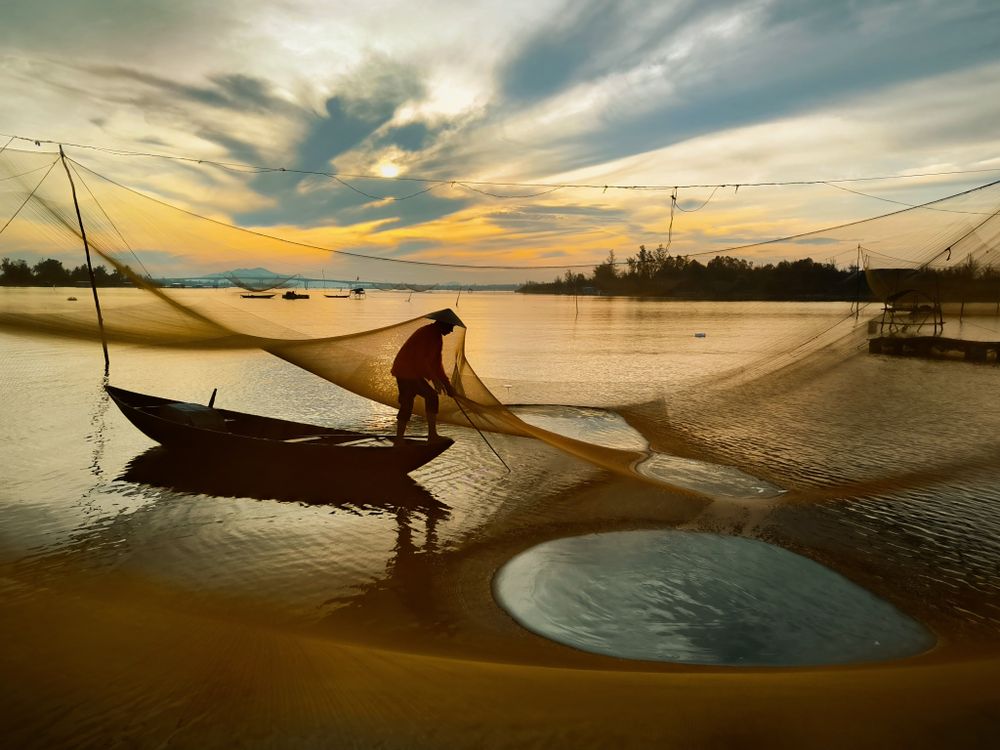 As the first light of day gently paints the horizon, a quiet silhouette appears upon the water. The fisherman casts his net into the cool, hope-filled morning, each knot carrying the wisdom of the past, each pull holding the promise of a new beginning. The wind whispers morning prayers through the threads, while the water cradles the golden and blue hues of the sky. This moment is the silent song of a waking world — the purest form of labor, patience, and the timeless bond between humankind and nature.