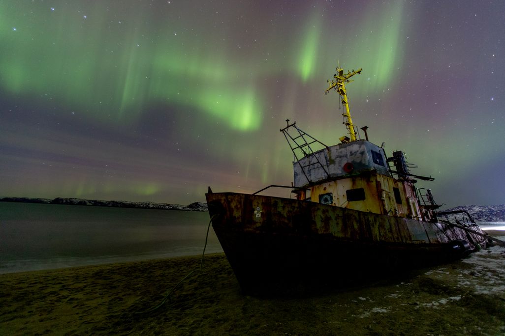 An image of a the northern lights in the sky. The photo also shows a dilapidated ship sitting on land next to water.