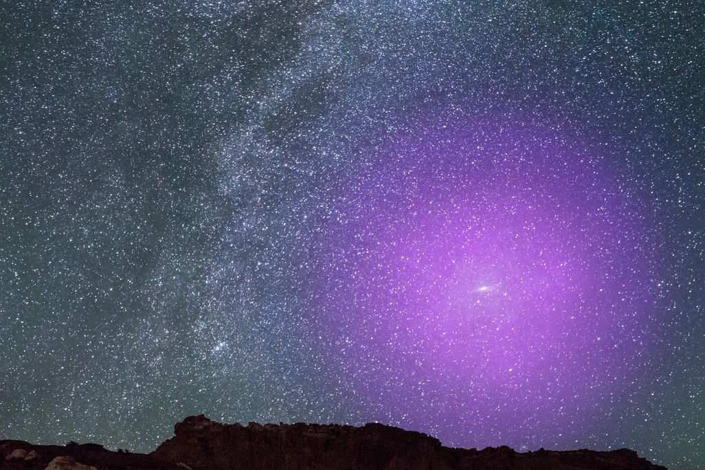 A view of rocks in foreground with an expansive, glittering night sky behind -- many stars and the curve of the Milky Way appear; in the lower righthand section of sky, an illustrated large circle of purple light indicates where Andromeda's halo would be