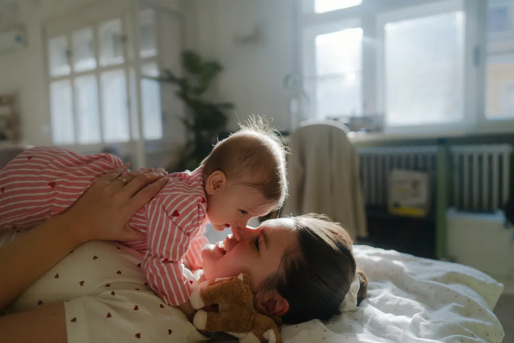 a woman lies on a bed holding her baby to her face, touching noses