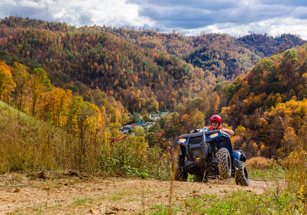 An off-road vehicle travels up a dirt trail at Burning Rock Off-Road Park, with rolling hills covered in peak fall foliage stretching across the background. Shades of gold, orange and red fill the forested landscape beneath a dramatic, cloud-filled sky.