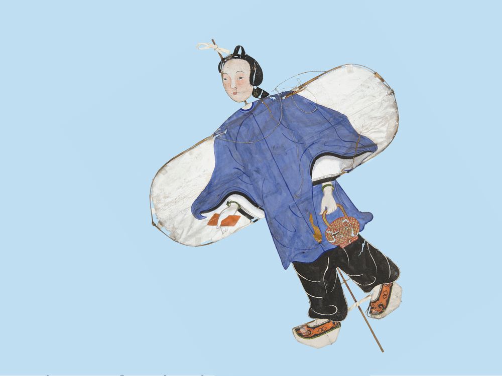 This paper kite depicts a Chinese girl wearing a long-sleeve blue tunic and loose-fitting black plants. A bamboo frame runs the length of the kite behind the paper.
