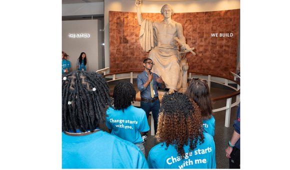 A group of students in bright blue shirts gather around a statue of George Washington with a museum facilitator