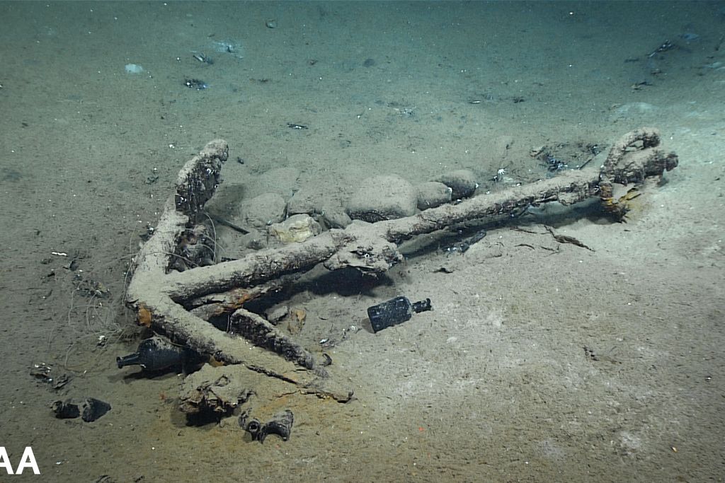 An anchor surrounded by bottles and other debris, covered in ocean sand and muck