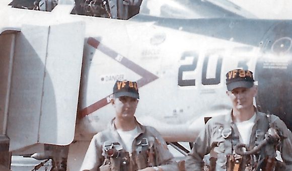 Two U.S. Navy men in uniform--a pilot and an aviator — stand in front of the cockpit of an F-4B Phantom II military jet aircraft.