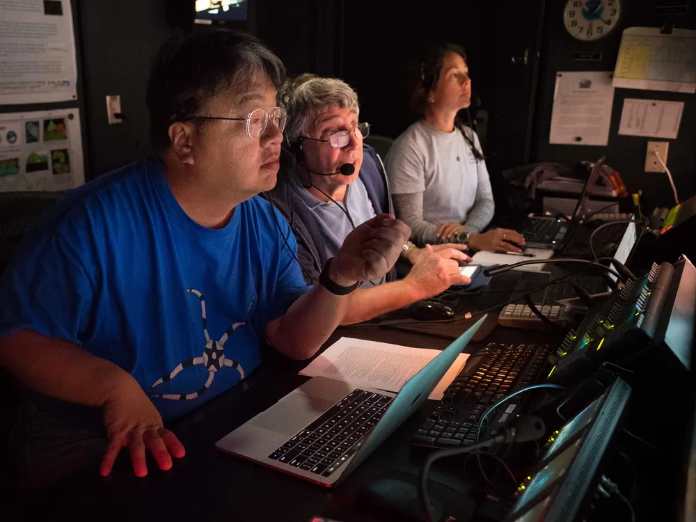 Onboard science leads Drs. Chris Mah and Chris Kelley and NOAA Educational Partnership Program Intern Nikola Rodriguez discuss and take a closer look at deepwater habitats explored with remotely operated vehicle Deep Discoverer on Horizon Guyot. (Image courtesy of the NOAA Office of Ocean Exploration and Research, 2017 Laulima O Ka Moana)