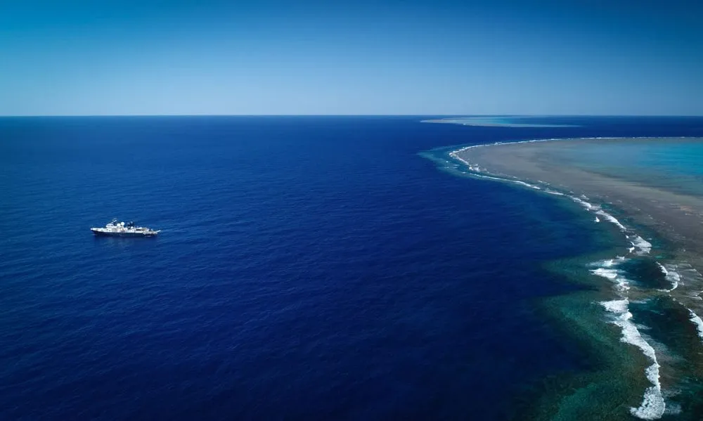 A photo taken from high above (likely by drone) shows the vast, bright blue ocean. The research vessel is on the left side. A long green and beige stretch of shallow coral reefs is on the right side. The horizon, where the deep blue ocean meets the lighte