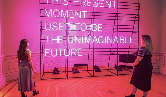 Two people stand in front of a pink neon sign that states "This Present Moment Used to Be the Unimaginable Future"