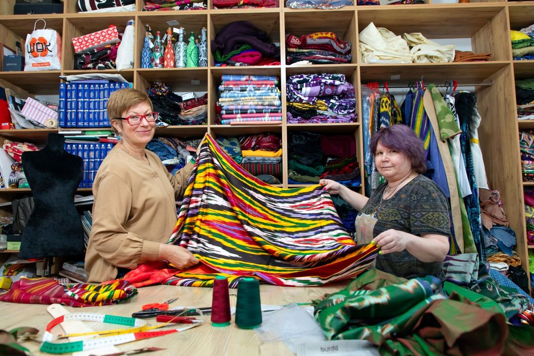 Lola Sayfi and another adult hold colorful fabric while looking at the camera and smiling. They stand behind a table and in front of shelves filled with crafts and clothing.