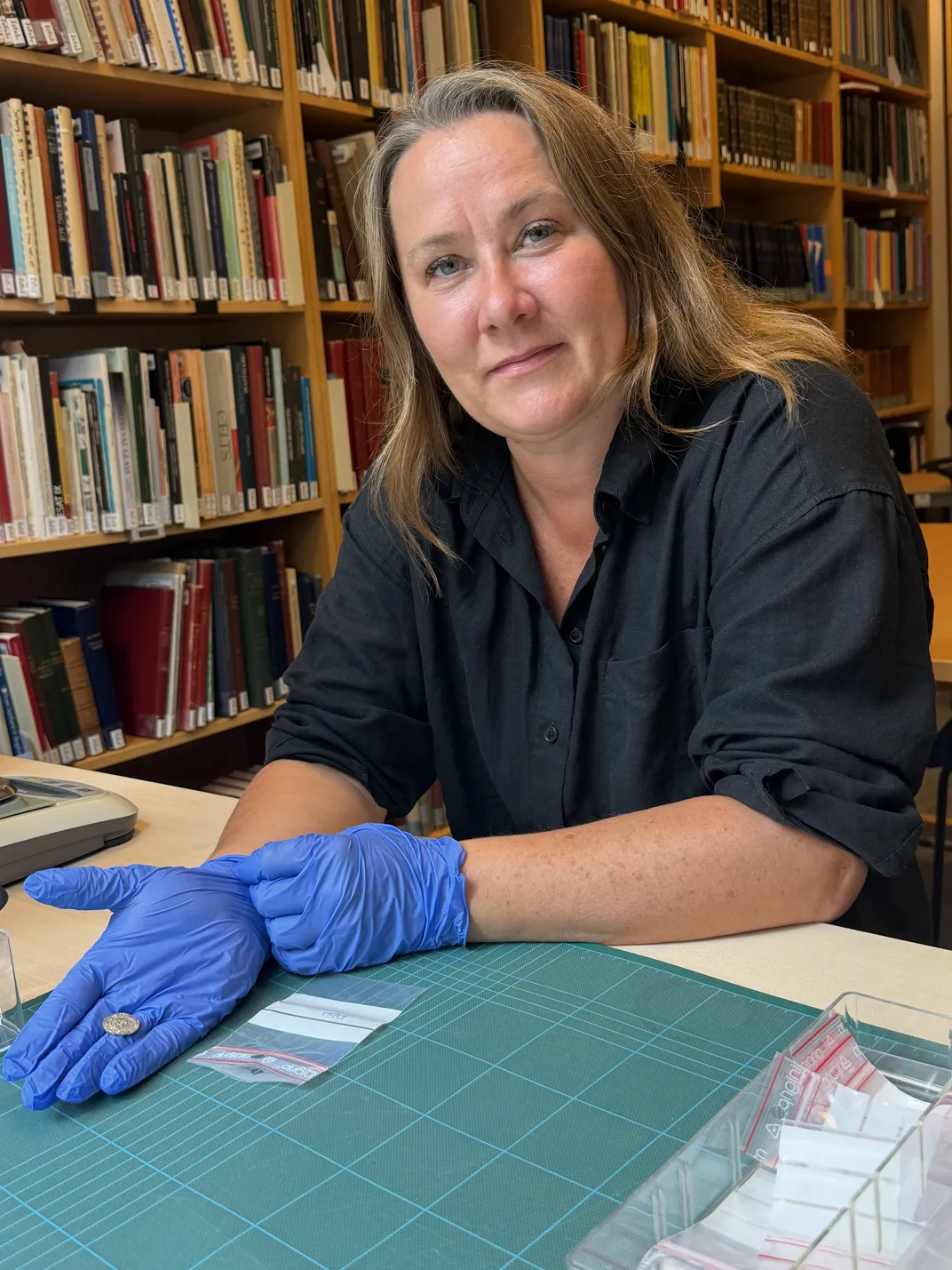 A woman wearing blue gloves sitting in a library holding a coin in one hand