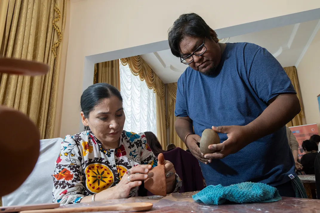 A woman, seated, and a man, standing next to her, both shape small clay bowls.