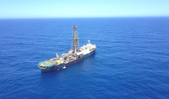 A large research vessel with a cell tower on top sails across a dark blue stretch of ocean that expands towards a paler blue sky along the horizon.