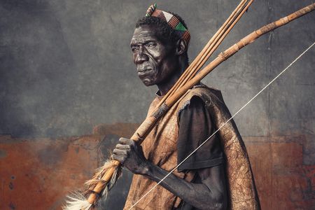 A Hadza elder wears a roughly tanned wild-animal skin over a T-shirt. The skin strips on his bow reinforce his weapon while the furs attest to his recent kills. His headband is not traditionally Hadza; members of the tribe have begun to adopt styles from neighboring groups.